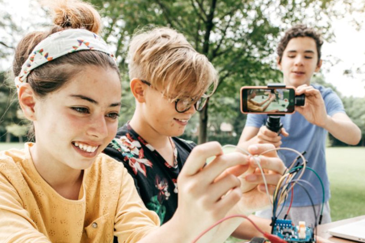 Leerlingen werken samen aan een technisch project buiten; één filmt met een smartphone terwijl de anderen experimenteren met draden en sensoren — een voorbeeld van leren door doen bij Atel!er Het Gooi.
